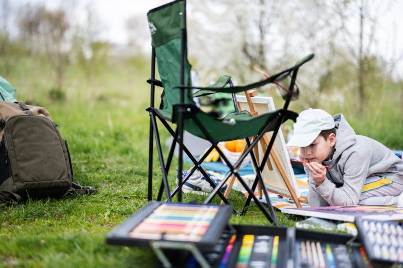 Boy enjoying outdoor on picnic blanket and painting on canvas while camping
