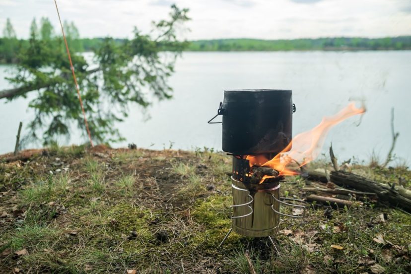 Camping wood stove cooking food stands on a metal, portable burner from which there is a flame, from the burning wood, on a rock, against the background of a lake and sky, on a summer evening.