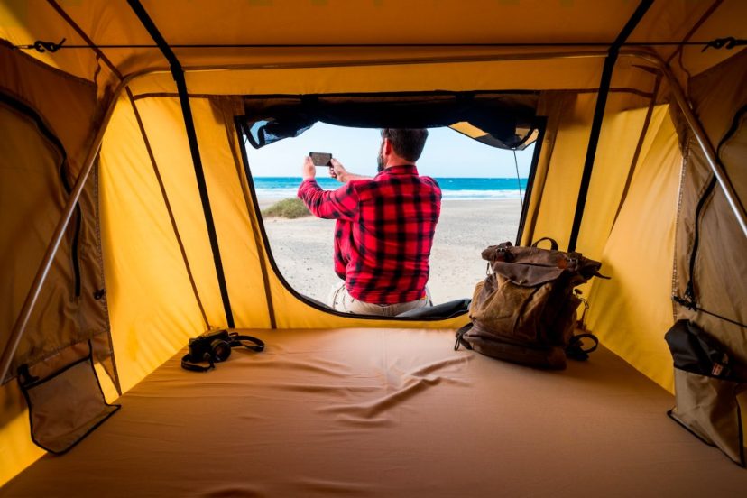 Freedom traveler man outside his SUV with camping tent in alternative vacation lifestyle in free camping at the beach taking picture of the natura landscape - backpack and camera inside and blue sea horizon in background