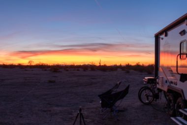 A blue truck is parked alongside a Wolf Pup Camper in an idyllic rural setting surrounded by lush greenery