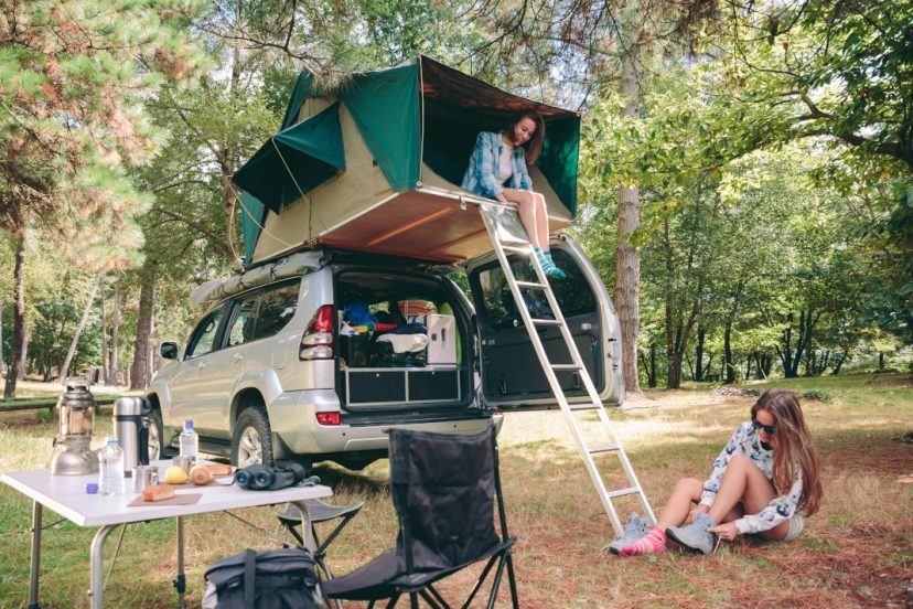 Young happy woman resting in a tent on the roof of SUV while other woman putting hiking boots sitting on the grass