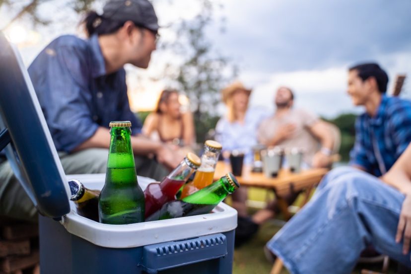 Group of friend having outdoors camping party together with camping fridge. Attractive young man and woman traveler drinking alcohol beer, enjoy hanging out during holiday vacation trip in forest.
