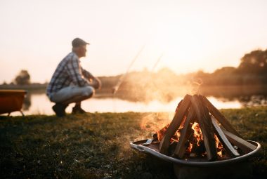 Close-up of campfire at sunset with man fishing in the background.